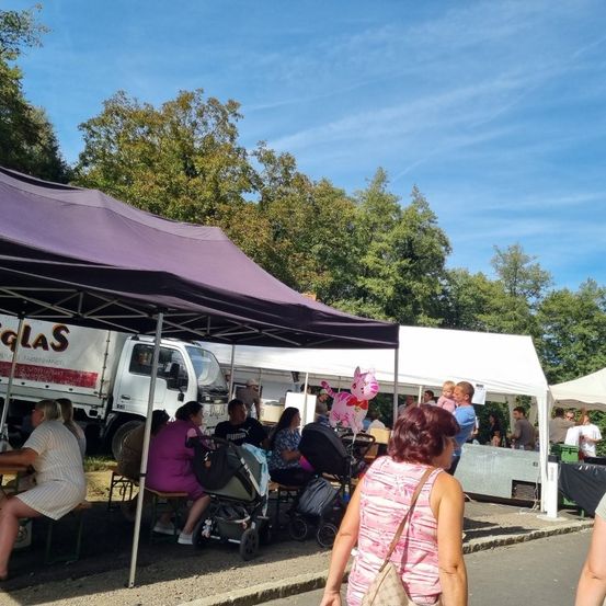 Eine Marktszene unter einem lilafarbenen Zelt mit verschiedenen Ständen. Menschen sitzen und stehen, einige mit Kinderwagen. Ein LKW ist dahinter geparkt und Bäume sind im Hintergrund.
