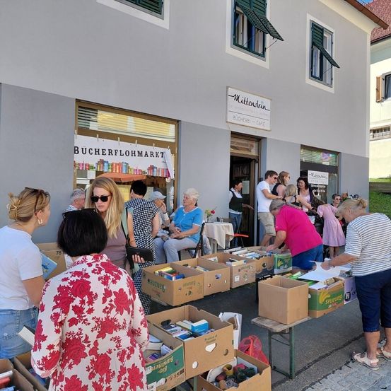 Vor einem Gebäude mit 'BUCHERFLOHMARKT' über dem Eingang findet ein Bücherflohmarkt statt. Verschiedene Menschen betrachten die Bücher in Kisten und einige stöbern.