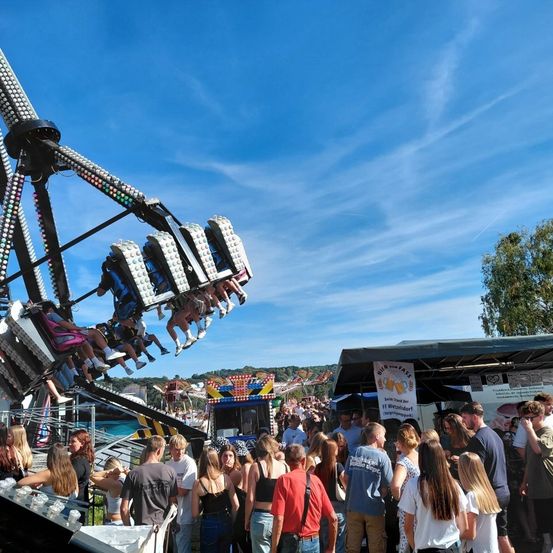 Eine große Menschenmenge versammelt sich in einem Vergnügungspark. Leute fahren auf einem Karussell und andere stehen herum. Ein Werbebanner ist auf einem Zelt im Hintergrund zu sehen.