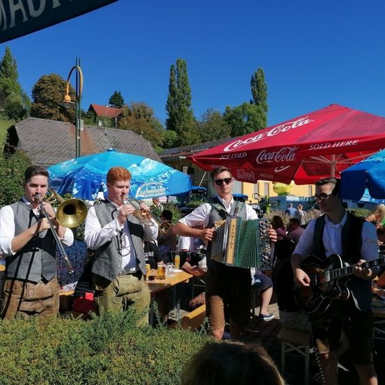 Eine Gruppe von Musikern spielt im Freien vor einem Publikum. Zwei Männer spielen Trompeten, einer spielt Akkordeon und der andere Gitarre. Hinter ihnen stehen bunte Schirme und Tische mit Getränken.