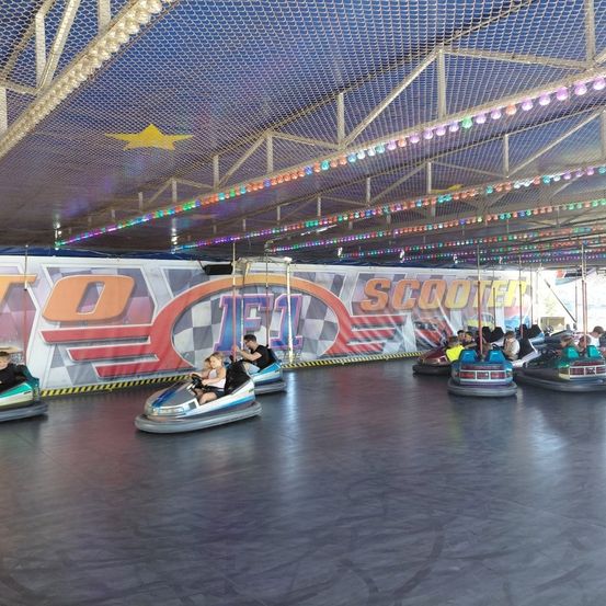 Eine Hüpfburg auf einem Jahrmarkt mit blauer Decke, gelbem Stern und bunten Lichtern. Leute in Bumper Cars mit einer Wand, die 'T0 JET' und 'SCOOTER' liest.