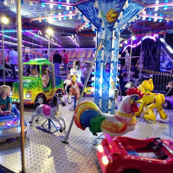 Ein bunter Karussell mit farbenfrohen Lichtern. Kinder fahren auf Spielzeugautos und Pferden. Einige sitzen auf einem grünen Lastwagen, andere auf einem roten Auto. Das Karussell ist mit blauen und lila Lichtern beleuchtet.
