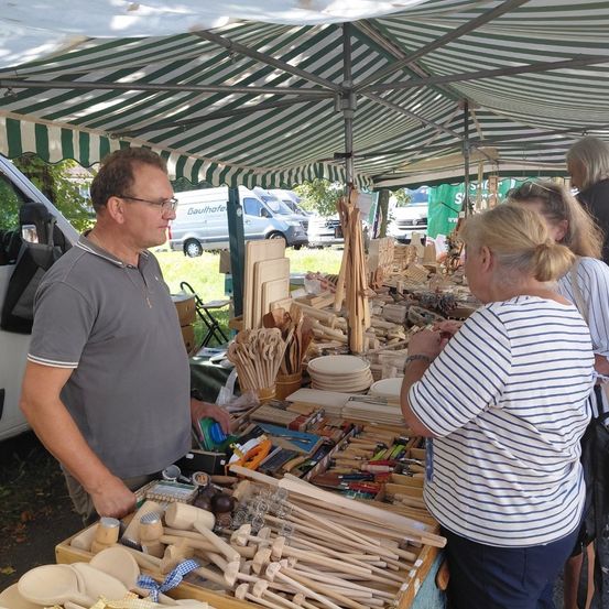 Ein Outdoor-Markt unter einem grün-weißen Zelt. Ein Mann und eine Frau schauen sich Holzartikel an. Im Hintergrund sind verschiedene Fahrzeuge geparkt.