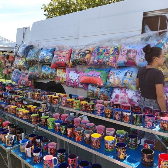 Ein Stand präsentiert verschiedene Tassen und Kissen mit Zeichentrickfiguren. Eine Frau steht neben dem Stand. Der Stand befindet sich in der Nähe eines weißen Lieferwagens.
