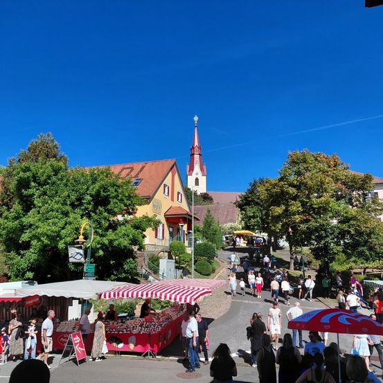 Eine Straßenszene mit einer Kirche, Geschäften und Fußgängern. Einige Geschäfte haben rote und weiße Markisen. Bäume säumen die Straße, und es gibt einen Turm mit einem Kirchturm.