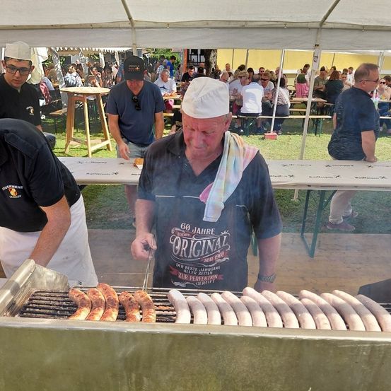 Zwei Männer grillen Würste unter einem Zelt, während andere an Picknicktischen sitzen. Ein Mann in einer Kochmütze bereitet Essen zu. Die Szene ist lebhaft, mit vielen anwesenden Personen.