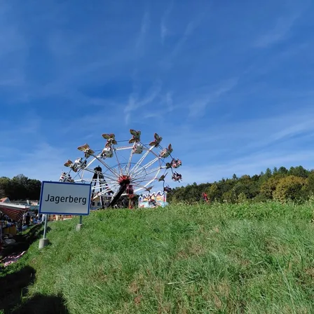Ein großes Riesenrad steht auf einem grasbewachsenen Hügel, mit einem Schild, das Jagerberg unten steht. Bäume und ein klarer blauer Himmel sind im Hintergrund.