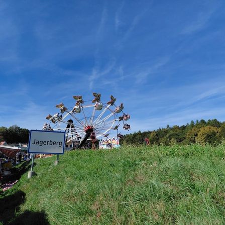 Ein großes Riesenrad steht auf einem grasbewachsenen Hügel, mit einem Schild, das Jagerberg unten steht. Bäume und ein klarer blauer Himmel sind im Hintergrund.
