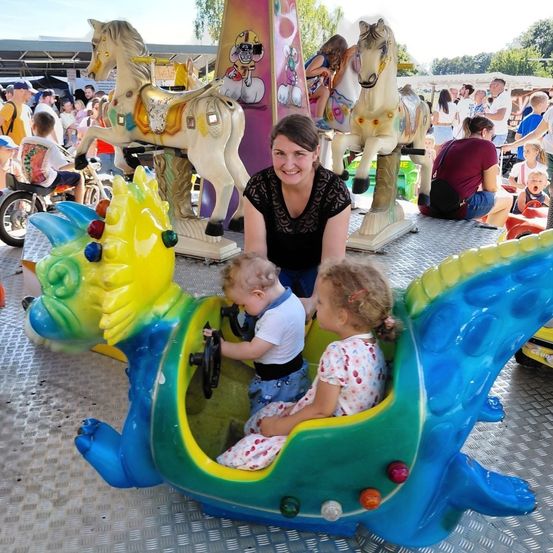Eine Frau mit zwei Kindern auf einer Fahrt in einem Vergnügungspark. Die Fahrt ist eine farbenfrohe, drachenförmige Karussell. Die Frau lächelt und hält die Hände der Kinder. Im Hintergrund sind Menschen und andere Fahrgeschäfte.