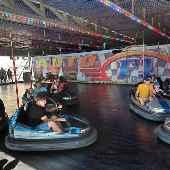 Eine Gruppe von Menschen fährt auf einer Indoor-Achterbahn. Die Autos sind blau und weiß. Bunte Lichter hängen von der Decke.