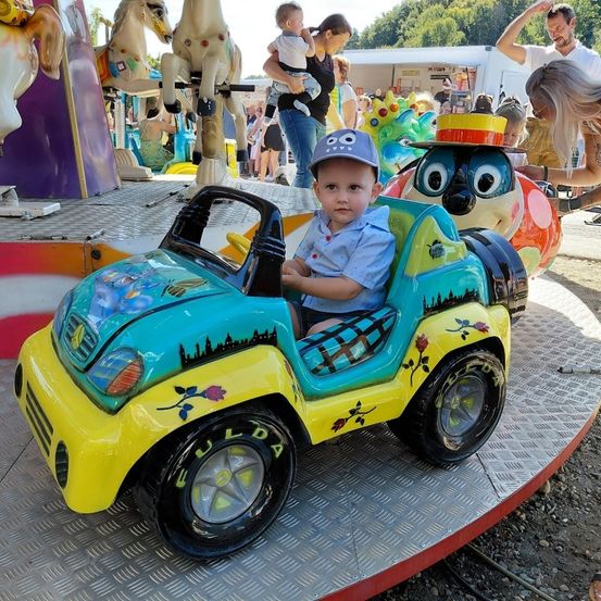 Ein kleines Kind fährt ein gelbes und blaues Spielzeugauto auf einem Jahrmarkt. Im Hintergrund sind andere Fahrgeschäfte zu sehen, darunter Pferde und ein Karussell.