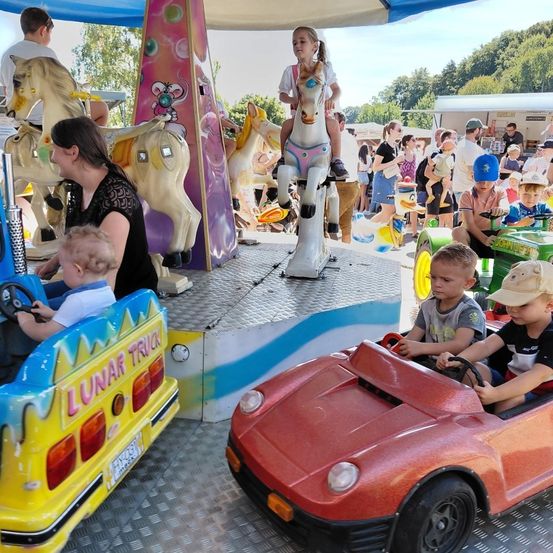 Auf einem Jahrmarkt beobachtet eine Frau ein Kind auf einem mechanischen Pferd. In der Nähe fahren Kinder in kleinen Autos. Der Jahrmarkt hat andere Fahrgeschäfte und Menschen im Hintergrund.