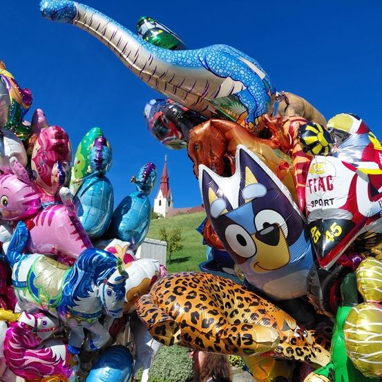 Eine farbenfrohe Sammlung bunter Luftballons mit verschiedenen Charakteren und Designs, in einem Haufen angeordnet, mit einer Kirchenspitze im Hintergrund unter einem klaren blauen Himmel.