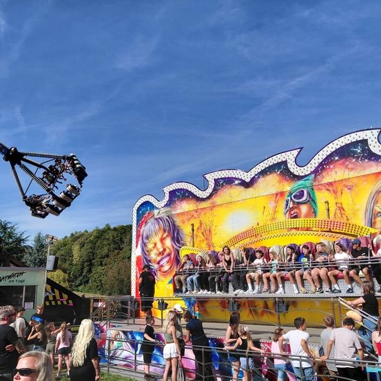 Eine Gruppe von Menschen genießt Fahrgeschäfte in einem Vergnügungspark. Eines der Fahrgeschäfte ist ein farbenfrohes Riesenrad, während andere auf einem Drehkarussell sitzen. Unten stehen Zuschauer.