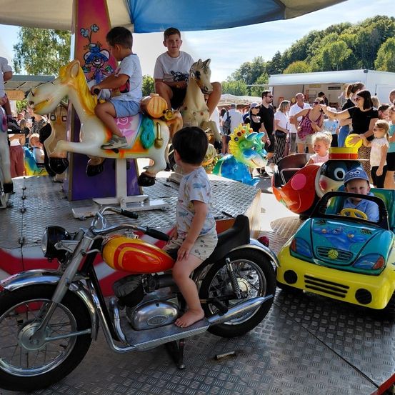Kinder reiten auf verschiedenen Fahrgeschäften im Vergnügungspark, darunter ein Pferd, ein Motorrad und ein Auto, unter einem Baldachin, während Zuschauer im Hintergrund stehen.