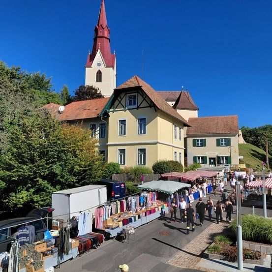 Eine geschäftige Marktszene vor einer Kirche mit einem Kirchturm. Verschiedene Stände präsentieren Kleidung und Gegenstände, viele Menschen gehen herum. Bäume und geparkte Autos sind im Bereich sichtbar.