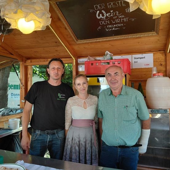 Drei Menschen stehen in einem kleinen Weinladen. Zwei Männer und eine Frau. Sie lächeln. Das Schild sagt: 'Der beste Wein ist mit Freunden.' Es gibt Flaschen und einen Kühler.