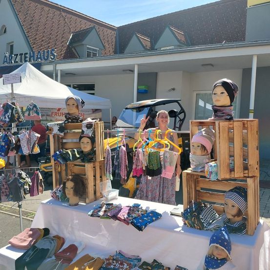 Eine Frau steht hinter einem Tisch mit verschiedenen Kleidungsstücken, darunter Hüte und Schals. Mannequins sind auf dem Tisch und auf Holzkisten platziert. Ein Gebäude mit dem Namen Arztehaus ist im Hintergrund.