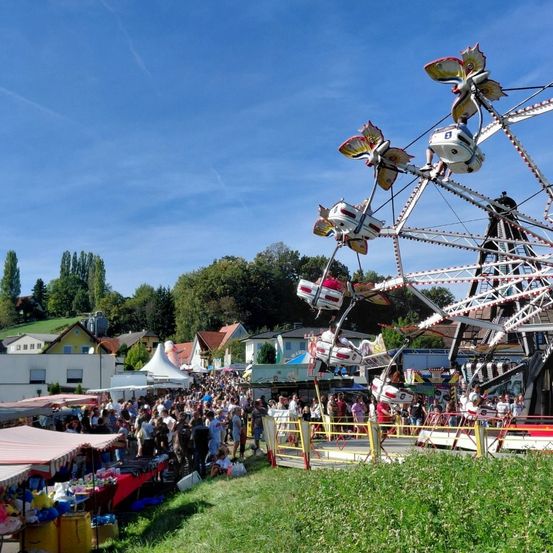 Ein lebendiges Jahrmarkt mit einem Riesenrad, einer Menschenmenge, Zelten und Ständen unter einem blauen Himmel mit Bäumen und Häusern im Hintergrund.