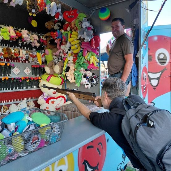 Zwei Männer an einem Karussellspielstand, einer schießt mit einer Luftgewehrpistole, während der andere zusieht, umgeben von hängenden Kuscheltieren.