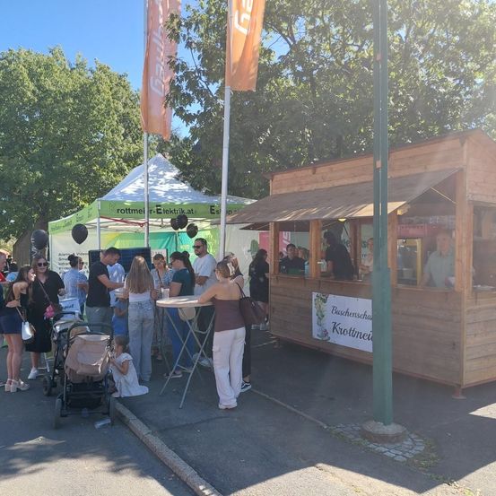 Eine Gruppe von Menschen versammelt sich draußen in der Nähe eines kleinen Holzhäuschens, möglicherweise auf einem Markt, mit mehreren Zelten und Fahnen im Hintergrund.
