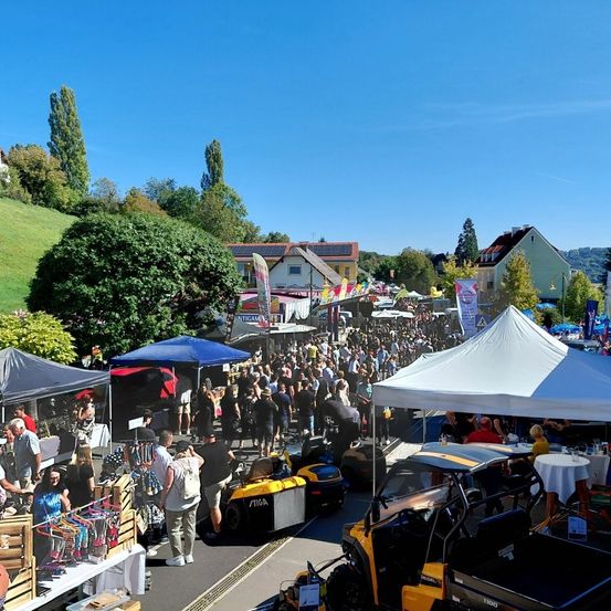 Eine überfüllte Veranstaltung im Freien zeigt eine Vielzahl von Zelten, Fahrzeugen und Menschen in einer sonnigen, lebendigen Umgebung mit üppigem Grün und Häusern im Hintergrund.