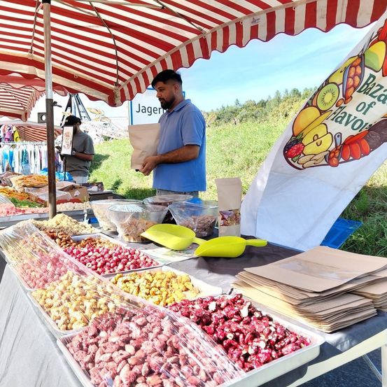 Ein Lebensmittelstand unter einem rot-weiß gestreiften Zelt präsentiert verschiedene Nüsse und Früchte. Ein Mann liest ein Papier und eine weitere Person steht hinter dem Stand.