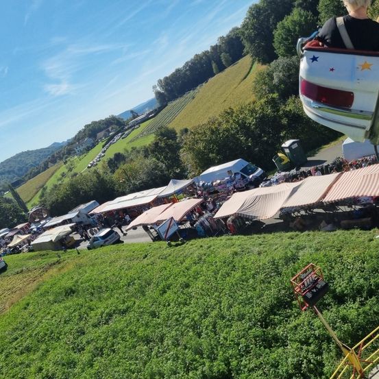 Luftaufnahme eines ländlichen Gebiets mit einem Jahrmarkt, Zelten und geparkten Autos. Eine Person fährt eine Karussell im Vordergrund. Bäume und Berge sind im Hintergrund.