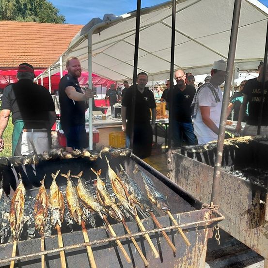 Mehrere Menschen sind um einen Grill versammelt, auf dem gekochte Fische liegen. Ein Mann steht lächelnd hinter dem Grill. Einige Leute tragen Hüte und Brillen. Im Hintergrund sind Zelte zu sehen.
