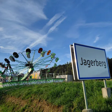 Ein Schild für Jagerberg steht auf einem Grashügel, mit einem bunten Riesenrad im Hintergrund.
