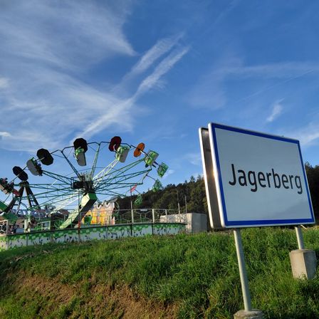 Ein Schild für Jagerberg steht auf einem Grashügel, mit einem bunten Riesenrad im Hintergrund.
