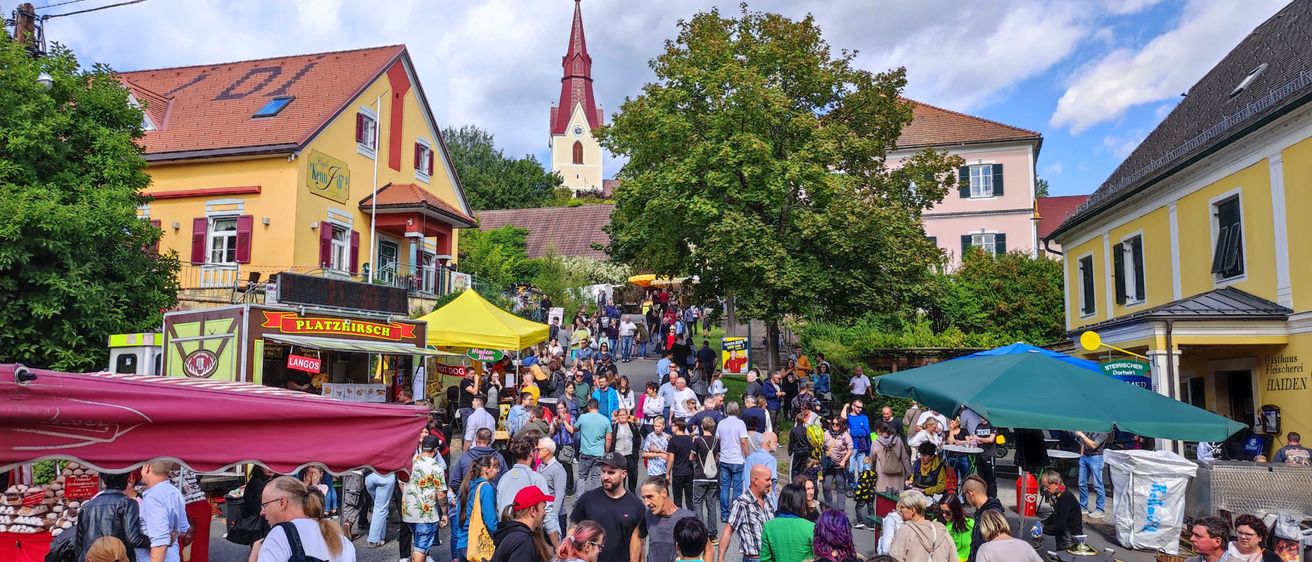Eine Menschenmenge versammelt sich auf einem Stadtplatz, mit Imbissständen und einer Kirche im Hintergrund.