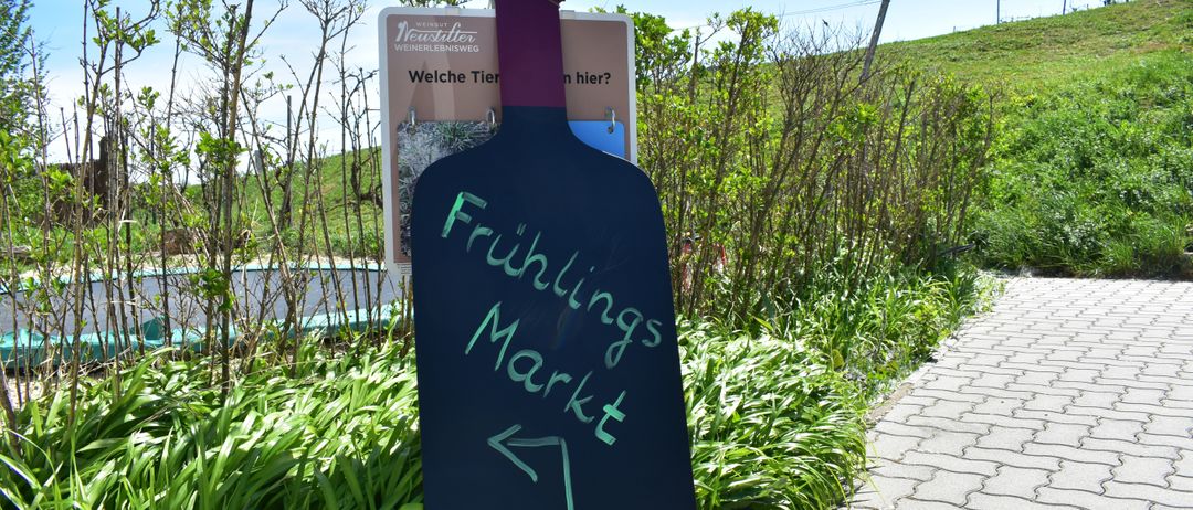 Ein Kreidebrett mit einer Flaschensilhouette zeigt auf einen Markt. Dahinter steht ein Schild mit 'Welche Tier'. Grüne Pflanzen und ein Weg führen zum Hügel.