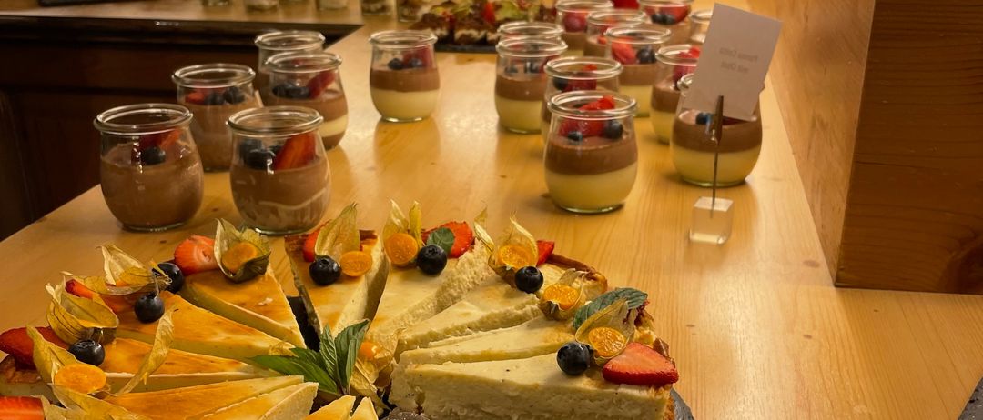Ein Holztisch hat einen geschnittenen Kuchen mit verschiedenen Früchten und Beeren. Dahinter stehen mehrere Desserts in Gläsern und ein Teller mit Essen.