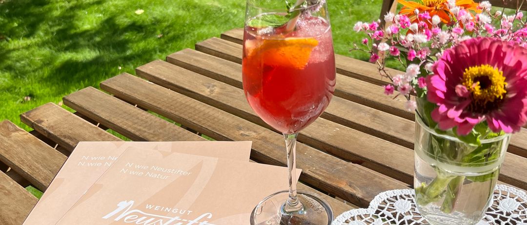Ein Tisch mit einem Glas rotem Cocktail, einem Blumenvase und einer Menükarte an einem sonnigen Tag.