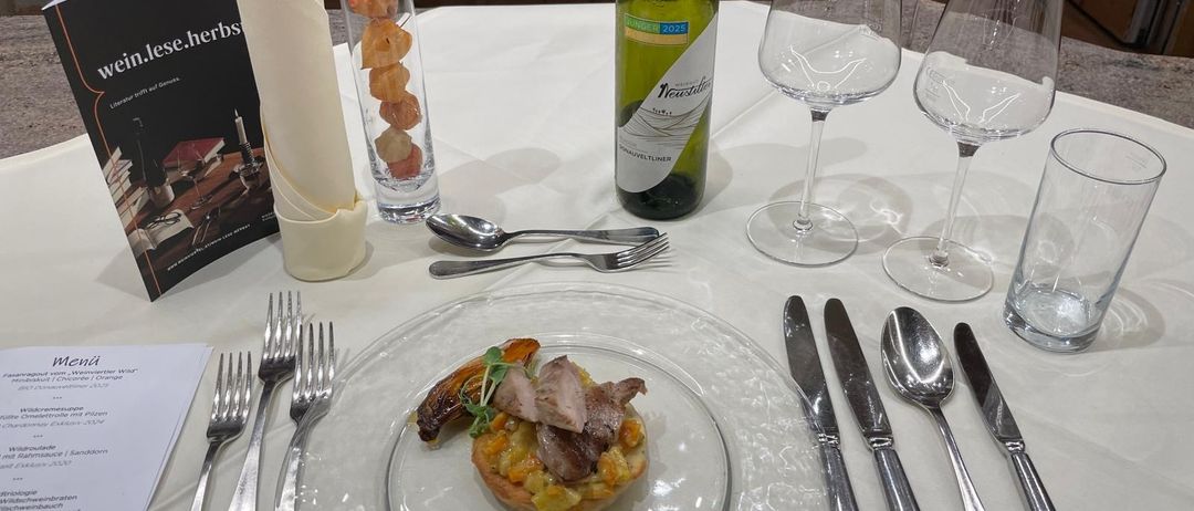 Ein Tisch mit einer Fleischplatte, einer Weinflasche und verschiedenen Besteckteilen. Die Platte enthält ein kleines Fleischgericht mit Gemüse.