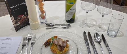 Ein Tisch mit einer Fleischplatte, einer Weinflasche und verschiedenen Besteckteilen. Die Platte enthält ein kleines Fleischgericht mit Gemüse.