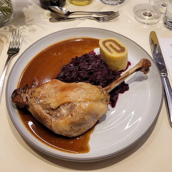Ein Teller mit einem gebratenen Entenschenkel, brauner Soße, Rotkohl und einer kleinen Teigrolle auf einer weißen Tischdecke.