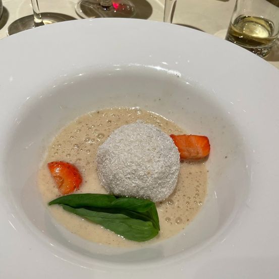 Ein weißer Teller mit einem Dessert bestehend aus einer runden weißen Kugel, einer cremigen Basis und Orangenscheiben. Eine grüne Blatt liegt neben dem Dessert.