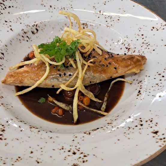 Ein Gourmetgericht mit einem knusprigen Teig, gefüllt mit Fleisch, garniert mit Petersilie und frittierten Nudeln, begleitet von einer reichhaltigen braunen Sauce und schwarzem Pfeffer auf einem weißen Teller mit braunen Sprenkeln.