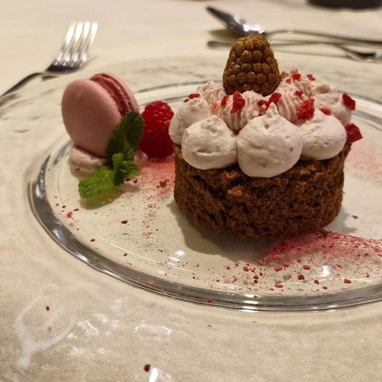 Ein kleiner Schokoladenkuchen mit Schlagsahne und roten Beeren auf einem Glasteller, begleitet von einem rosa Macaron und einem Minzblatt.
