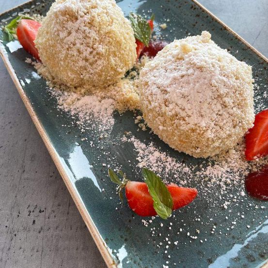 Zwei kugelförmige Desserts, mit Puderzucker bestreut, auf einem türkisfarbenen Teller mit Erdbeeren und Minzblättern.