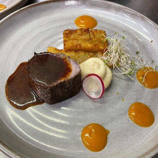 Ein Gourmetgericht auf einem Teller mit einem Stück Fleisch in brauner Sauce, Kartoffelpüree, frittierten Kartoffeln und Radieschen mit Sprossen und orangefarbener Sauce.