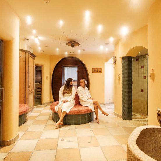 Zwei Frauen in weißen Roben sitzen auf einer runden Bank in einem Spa, mit gefliestem Boden und gelben Wänden.