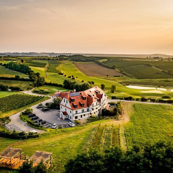 Eine Luftaufnahme eines großen weißen Gebäudes mit roten Dächern und Solarmodulen, umgeben von grünen Feldern und einem Golfplatz.