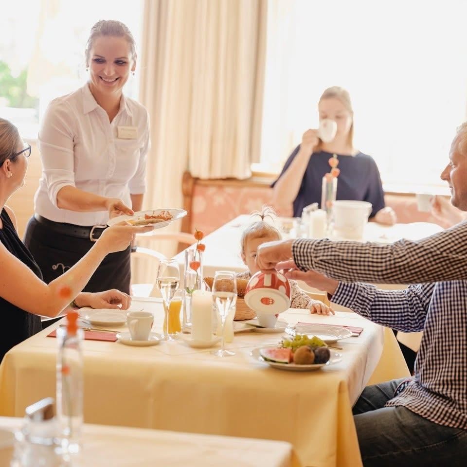 A family is having breakfast at a restaurant. The waiter is serving food to the family. The baby is drinking from a cup.