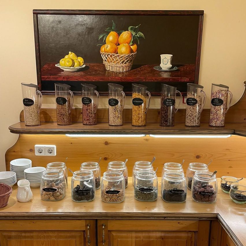 A wooden counter with various glass jars filled with different grains and seeds. Above, a wooden shelf holds more jars and a painting of lemons and oranges.