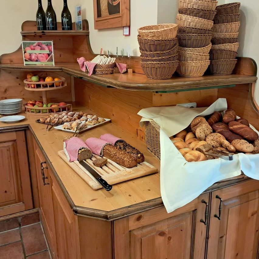 A wooden counter in a bakery displays various breads, pastries, and fruits. Wicker baskets hold more bread, and bottles of wine are on a shelf.