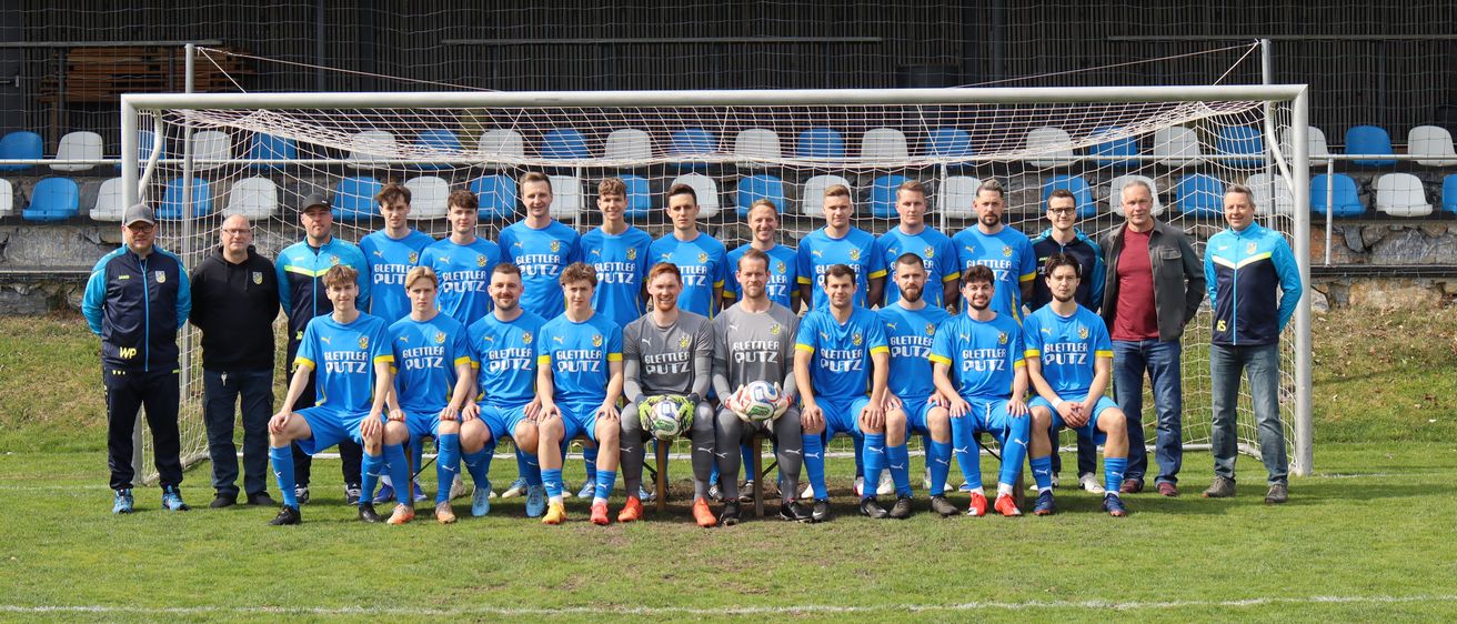 Eine Fußballmannschaft in blauen Trikots posiert für ein Foto auf einem Feld mit einem Tor im Hintergrund.