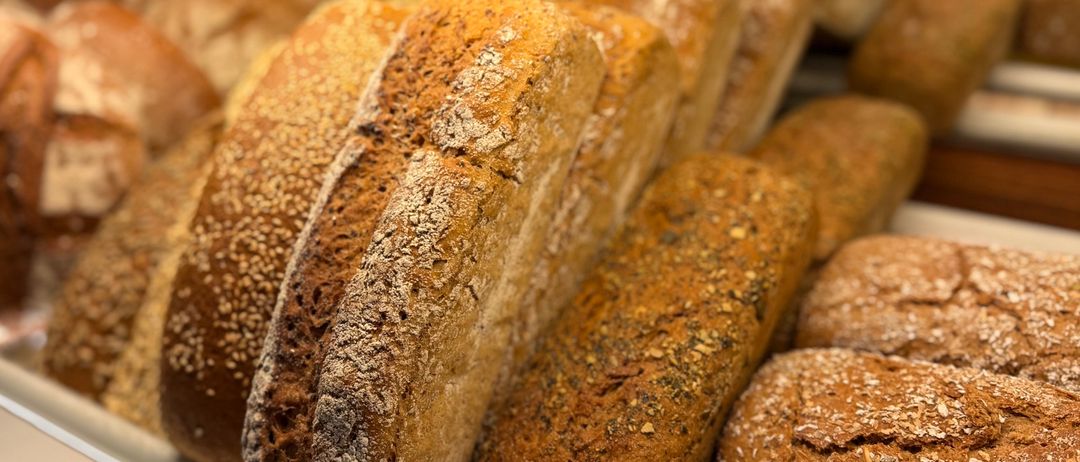 Eine Bäckerei-Auslage mit verschiedenen Brotsorten, darunter Vollkorn-, Roggen- und Mischbrote. Jedes Brot ist mit Samen oder Körnern bestreut und ordentlich auf einem weißen Regal angeordnet.
